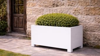 Donica aluminiowa Apollo 18 Mobile stoi na kamiennym patio przy ścianie i drewnianych drzwiach, trzymając okrągły zielony krzew. Światło słoneczne rzuca delikatne cienie, podkreślając czysty, minimalistyczny design donicy.