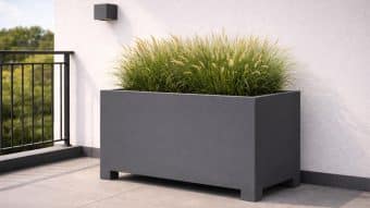 Nowoczesna szara Donica aluminiowa Apollo 18 (60 cm wysokości) wypełniona wysokimi zielonymi trawami ozdobnymi stoi na wyłożonym kafelkami balkonie przy białej ścianie. Po lewej stronie znajduje się czarna balustrada, za nią rozmywają się zielone drzewa, a powyżej zamontowana jest kwadratowa lampa ścienna.