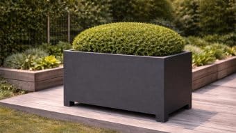 Duża, prostokątna, matowo-czarna Donica aluminiowa Apollo 19 Mobile stoi na drewnianym tarasie, trzymając okrągły krzew. Za nim znajdują się podniesione grządki z zielonymi liśćmi i druciane ogrodzenie pośród bujnej zieleni.