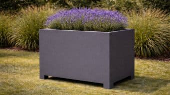 Donica aluminiowa Apollo 19, prostokątna ciemnoszara donica o krótkich nogach i nowoczesnych liniach, stoi na trawie wypełnionej kwitnącą lawendą. Bujne trawy ozdobne tworzą zielone tło.