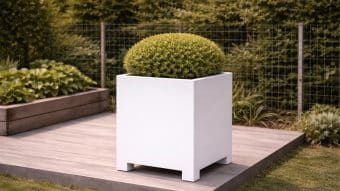 Okrągły, starannie przycięty zielony krzew znajduje się w dużej, białej donicy aluminiowej Apollo 2 Mobile na drewnianym tarasie z bujną roślinnością i drucianym ogrodzeniem w tle - prezentując dobrze utrzymany ogród lub patio.