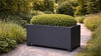 Donica aluminiowa Apollo 20 Mobile, duża prostokątna ciemnoszara donica, stoi na żwirowej ścieżce ogrodowej z gęstym, okrągłym krzewem. Bujne zielone rośliny i drzewa otaczają ścieżkę, tworząc spokojną scenerię na zewnątrz.