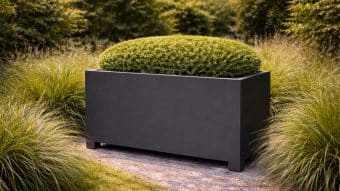 Donica aluminiowa Apollo 21 Mobile to starannie przycięty zielony krzew o owalnym kształcie. Stoi na żwirowej ścieżce pośród ozdobnych traw i bujnej zieleni w jasnym, zadbanym ogrodzie.