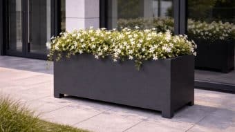 Duży, prostokątny Donica aluminiowa Apollo 22 Mobile wypełniony kwitnącymi białymi kwiatami i bujną zielenią stoi na wyłożonym kafelkami patio przed szklanymi drzwiami i oknami, a liście rozlewają się po krawędziach i odbijają w szkle.