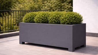 Na wyłożonym kafelkami patio stoi Donica aluminiowa Apollo 22, duża i matowo szara, trzymająca starannie przycięte okrągłe zielone krzewy. W tle czarna metalowa balustrada i zielone drzewa otaczają część białej ściany po prawej stronie.