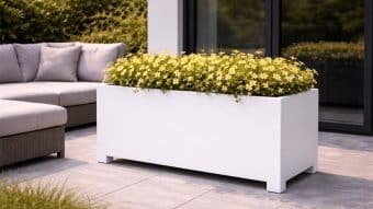 Nowoczesne patio prezentuje Donicę aluminiową Apollo 23 Mobile wypełnioną żółtymi kwiatami, obok szarej sofy zewnętrznej na szarych płytkach. Szklane drzwi i bujne zielone krzewy dodają przestrzeni elegancji.