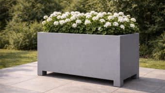 Donica aluminiowa Apollo 23, duża prostokątna donica aluminiowa, stoi na wyłożonym kafelkami patio wypełnionym bujną zielenią i białymi kwiatami, podczas gdy gęste krzewy i drzewa tworzą naturalne tło.