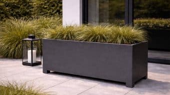 Donica aluminiowa Apollo 24 Mobile, wypełniona wysokimi trawami ozdobnymi, stoi na wyłożonym kafelkami patio przy dużym oknie. Czarna metalowa latarnia z białą świecą obok bujnych krzewów dopełnia tę spokojną aranżację.