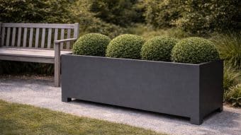 Elegancka Donica aluminiowa Apollo 25 Mobile prezentuje cztery starannie przycięte, okrągłe, zielone krzewy i stoi na żwirowej ścieżce obok drewnianej ławki, z bujną zielenią i trawą w tle, tworząc schludny, zachęcający wygląd ogrodu.