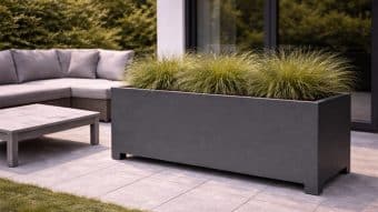 Na nowoczesnym patio znajduje się duża Donica aluminiowa Apollo 26 Mobile z wysokimi trawami ozdobnymi na jasnoszarych płytkach. W pobliżu, obok szklanych drzwi i bujnych zielonych żywopłotów, znajduje się szara sofa segmentowa z poduszkami i pasujący do niej niski stolik kawowy.