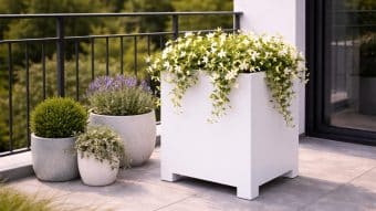 Na nowoczesnym patio znajduje się Donica aluminiowa Apollo 3 Mobile o zwisających białych kwiatach, uzupełniona trzema okrągłymi doniczkami z lawendą, krzaczastą zielenią i srebrzystymi liśćmi. Przestrzeń jest jasna i otoczona czarną balustradą oraz bujnymi drzewami.