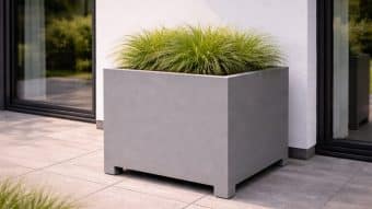 Duża, nowoczesna Donica aluminiowa Apollo 6 Mobile z wysoką trawą ozdobną stoi na jasnoszarym patio wyłożonym kafelkami obok białej ściany i szklanych drzwi, odbijając zieloną scenerię na zewnątrz.