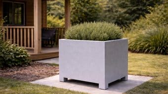 Duża, kwadratowa, jasnoszara Donica aluminiowa Apollo 6 obsadzona gęstymi zielonymi krzewami stoi na wybrukowanym terenie ogrodu przy drewnianej werandzie, otoczona bujną trawą, roślinami ozdobnymi, drzewami i częścią drewnianego domu.