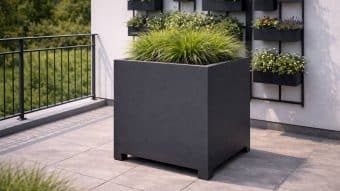 Duża, kwadratowa, matowo-czarna Donica aluminiowa Apollo 7 Mobile wypełniona wysoką, zieloną trawą ozdobną stoi na szarym, wyłożonym kafelkami tarasie. Za nim znajdują się zamontowane na ścianie czarne donice z kwitnącymi kwiatami. Taras otoczony jest metalową balustradą, a za nią rosną drzewa.