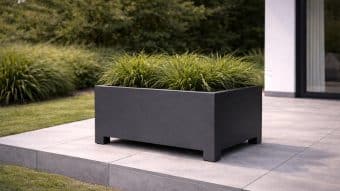 Nowoczesna Donica aluminiowa Apollo 8, mierząca 70 cm wysokości i wypełniona wysoką trawą ozdobną, stoi na szarym patio wyłożonym kafelkami w pobliżu nowoczesnego budynku z dużymi szklanymi drzwiami. W tle pojawia się bujna zieleń.