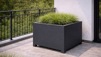 Duża Donica aluminiowa Apollo 9 Mobile w kolorze ciemnoszarym, wypełniona bujną zieloną trawą ozdobną, stoi na wyłożonym kafelkami balkonie obok białej ściany i czarnej balustrady, z gęstą zielenią widoczną na zewnątrz.