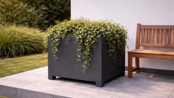 Donica aluminiowa Apollo 9 z bujnymi zielonymi kaskadowymi liśćmi znajduje się na wyłożonym kafelkami patio obok drewnianej ławki, z białą ścianą i trawą krajobrazową oraz krzewami w tle, tworząc spokojną scenerię na zewnątrz.