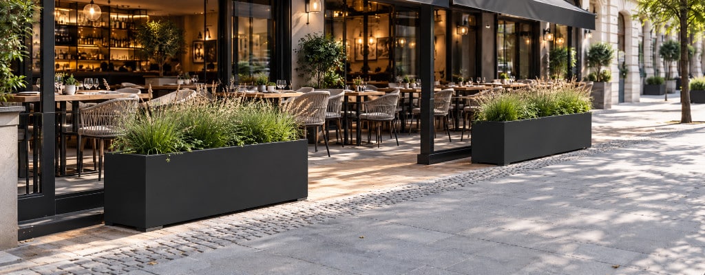 Dwie krótkie, czarne donice aluminiowe z trawami ozdobnymi, ustawione wzdłuż chodnika i oddzielające ogródek restauracyjny od przestrzeni dla pieszych.