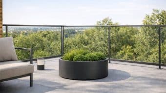 Nowoczesny balkon ze szklanymi balustradami wychodzi na bujny krajobraz. Na podłodze wyłożonej kafelkami znajduje się Donica aluminiowa Ada 2 wypełniona trawami ozdobnymi. Wyściełane krzesło i mała latarnia zewnętrzna dopełniają spokojne, minimalistyczne otoczenie.