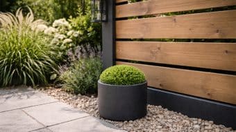 Okrągły, wypielęgnowany zielony krzew siedzi w Donica aluminiowa Carla 3 na małych kamykach obok drewnianego ogrodzenia. Trawy ozdobne, lawenda, białe rośliny kwitnące i czarna latarnia uzupełniają stylową scenę ogrodową.