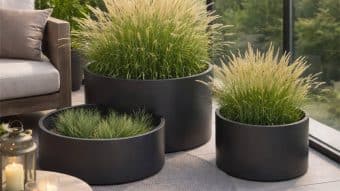 Zestaw Donica aluminiowa Daria 3 w kolorze czarnym, każdy na innej wysokości, prezentuje ozdobne trawy na patio z jasnymi płytkami. Beżowa sofa, świece, zielone drzewa i teksturowany dywan podkreślają przytulną, nowoczesną atmosferę na zewnątrz.