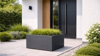 Stylowe wejście z eleganckimi czarnymi drzwiami, długą pionową klamką i lampą ścienną. Donica ogrodowa Alessio 12 prezentuje wysokie zielone trawy na jasnoszarym patio wyłożonym kafelkami, otoczonym wypielęgnowanymi krzewami i żwirem.