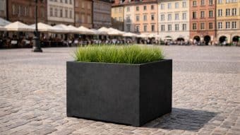 Duża czarna Donica ogrodowa Davide 4 wypełniona zieloną trawą stoi na brukowanym placu. Za nią kolorowe zabytkowe budynki, kawiarniane stoliki z parasolami i niewyraźne postacie w oddali.