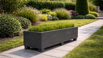 Prostokątna, ciemnoszara Donica ogrodowa Francesco 10 z wysoką zieloną trawą stoi na wybrukowanym chodniku obok wypielęgnowanego trawnika i bujnego, zagospodarowanego ogrodu z różnymi krzewami i trawami ozdobnymi w tle.