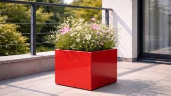 Donica ogrodowa Marco 1 w żywym czerwonym kolorze, wypełniona białymi stokrotkami i różowymi kosmosami, stoi na słonecznym tarasie wyłożonym jasnoszarymi płytkami, w pobliżu metalowej balustrady i z widokiem na zielone drzewa.