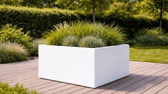 Donica ogrodowa Marco 10, duża nowoczesna biała donica wypełniona trawami ozdobnymi, stoi na drewnianym tarasie pośród bujnego ogrodu z drzewami i żywopłotami. Światło słoneczne rzuca delikatne cienie, podkreślając spokojną atmosferę na zewnątrz.