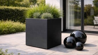 Nowoczesne patio prezentuje dużą donicę ogrodową Marco 11 wypełnioną trawami ozdobnymi i lawendą, wraz z trzema błyszczącymi czarnymi kulami dekoracyjnymi na wyłożonej kafelkami powierzchni. Tło dopełniają szklane drzwi i zielone krzewy.