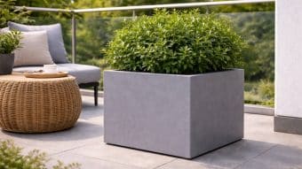 Duża kwadratowa Donica ogrodowa Marco 4 z bujnymi zielonymi roślinami stoi na nowoczesnym patio z szarymi płytkami. Za nim znajduje się wiklinowy stolik z filiżanką, wyściełane krzesło i szklana balustrada z widokiem na zieleń.