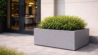 Donica ogrodowa Marco 6, wypełniona bujnymi zielonymi krzewami, stoi na wyłożonym kafelkami patio obok nowoczesnego budynku z przeszklonymi drzwiami i beżowymi ścianami. Przez drzwi widać stoliki i krzesła restauracji.