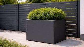 Duża, ciemnoszara donica ogrodowa Marco 7 podtrzymuje gęsty, zielony krzew. Donica stoi na jasnoszarym bruku przed nowoczesnym ogrodzeniem z czarnych listew, z jasnozielonymi liśćmi i drzewami w tle.