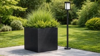 Donica ogrodowa Marco 9, wypełniona wysokimi zielonymi trawami i krzewami, stoi na kamiennym patio obok eleganckiej czarnej lampy zewnętrznej. Bujna trawa, krzewy i drzewa tworzą żywe tło w zadbanym ogrodzie.