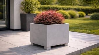 Kwadratowa biała donica ogrodowa Nicola 4 z krótkimi nóżkami eksponuje krzew o gęstych czerwonawych liściach na nowoczesnym patio z dużych szarych płytek. W tle znajduje się kolejna donica, zielony krzew i przycięty trawnik otoczony żywopłotem.