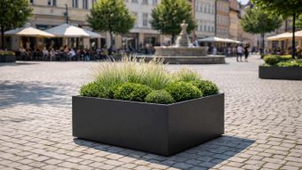 Duża donica Alessio 13, obsadzona ozdobnymi trawami i zielonymi krzewami, znajduje się na brukowanym placu wyłożonym drzewami. Ludzie relaksują się przy kawiarnianych stolikach pod parasolami w pobliżu, z budynkami i fontanną w tle.