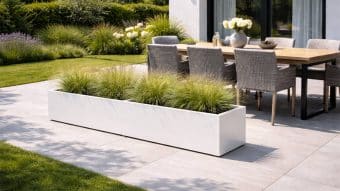 Nowoczesne patio obejmuje Dużą donicę Alessio 9 - długą białą prostokątną donicę z wysokimi trawami. W pobliżu drewniany stół z sześcioma plecionymi krzesłami i kwiatowymi elementami centralnymi znajduje się pośród bujnego trawnika i ogrodu.