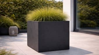 Duża donica Marco 8 w kolorze matowej czerni z trawą ozdobną stoi na jasnoszarym patio wyłożonym kafelkami obok dekoracyjnej latarni. Bujne zielone rośliny i szklane drzwi odbijają światło słoneczne, tworząc nowoczesną, świeżą przestrzeń zewnętrzną.