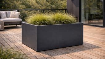 Duża, prostokątna, matowo czarna Duża donica Marco 9 wypełniona wysoką, zieloną trawą ozdobną stoi na drewnianym tarasie. Za nim znajduje się nowoczesna sofa zewnętrzna, szklane drzwi i drzewa widoczne za czarną balustradą.