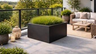 Nowoczesny balkon ze szklaną balustradą, na którym znajduje się Duża donica Alessio 7 wypełniona wysoką zieloną trawą, wiklinowe krzesła z beżowymi poduszkami, drewniany stół, rośliny doniczkowe, dywan i dekoracyjne lampiony. Na zewnątrz widoczne są drzewa i zieleń.