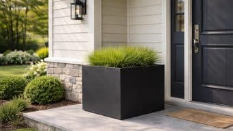 Duża donica Alessio 15 w eleganckiej czerni, wypełniona wysoką trawą ozdobną, stoi na szarym ganku przy ciemnych drzwiach wejściowych. Dom ma jasny siding, kamienne akcenty, latarnię ścienną, wycieraczkę i zagospodarowane podwórko z krzewami i zieloną trawą.
