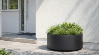 Duża donica Ada 1 stoi na nowoczesnym patio przy białym wejściu, wypełnionym zielonymi trawami ozdobnymi i liściastymi krzewami. Światło słoneczne rozjaśnia minimalistyczne, czyste otoczenie i rzuca miękkie cienie na ziemię.