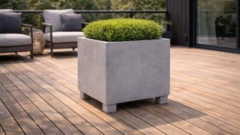 Duża donica Nicola 4, kwadratowa szara donica, prezentuje starannie przycięty okrągły krzew na drewnianym tarasie z szarymi wyściełanymi krzesłami, czarną metalową balustradą i zielenią w tle, tworząc nowoczesny wygląd na zewnątrz.