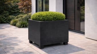 Nowoczesne patio obejmuje Dużą donicę Nicola 5, dużą matową czarną kwadratową donicę z okrągłym zielonym krzewem, ustawioną na szarych płytkach przy szklanych drzwiach i kontrastujących ścianach, otoczoną żywą zielenią.