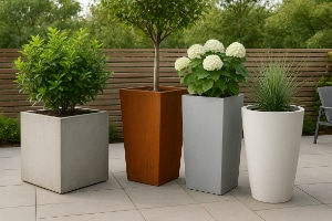Cztery nowoczesne donice na tarasie — szara kwadratowa z zielonym krzewem, wysoka rdzawa z małym drzewkiem, jasnoszara z białymi hortensjami oraz biała, wysoka donica z trawiastą rośliną; w tle drewniany płot i zieleń drzew.