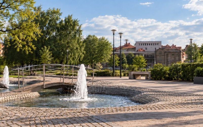 Czarna donica Geraldo 2 przy miejskiej fontannie Czarna betonowa donica Geraldo 2 z cisem przy fontannie w miejskim parku