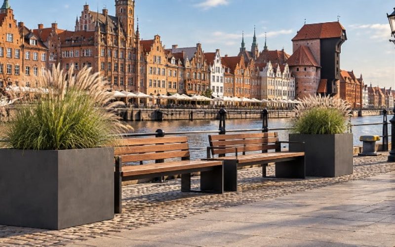 Donice Monica z trawami i ławkami na nadwodnej promenadzie w Gdańsku Dwie grafitowe donice Monica z trawą ozdobną ustawione przy nabrzeżu w Gdańsku, z ławkami pomiędzy nimi i widokiem na historyczną zabudowę portową.